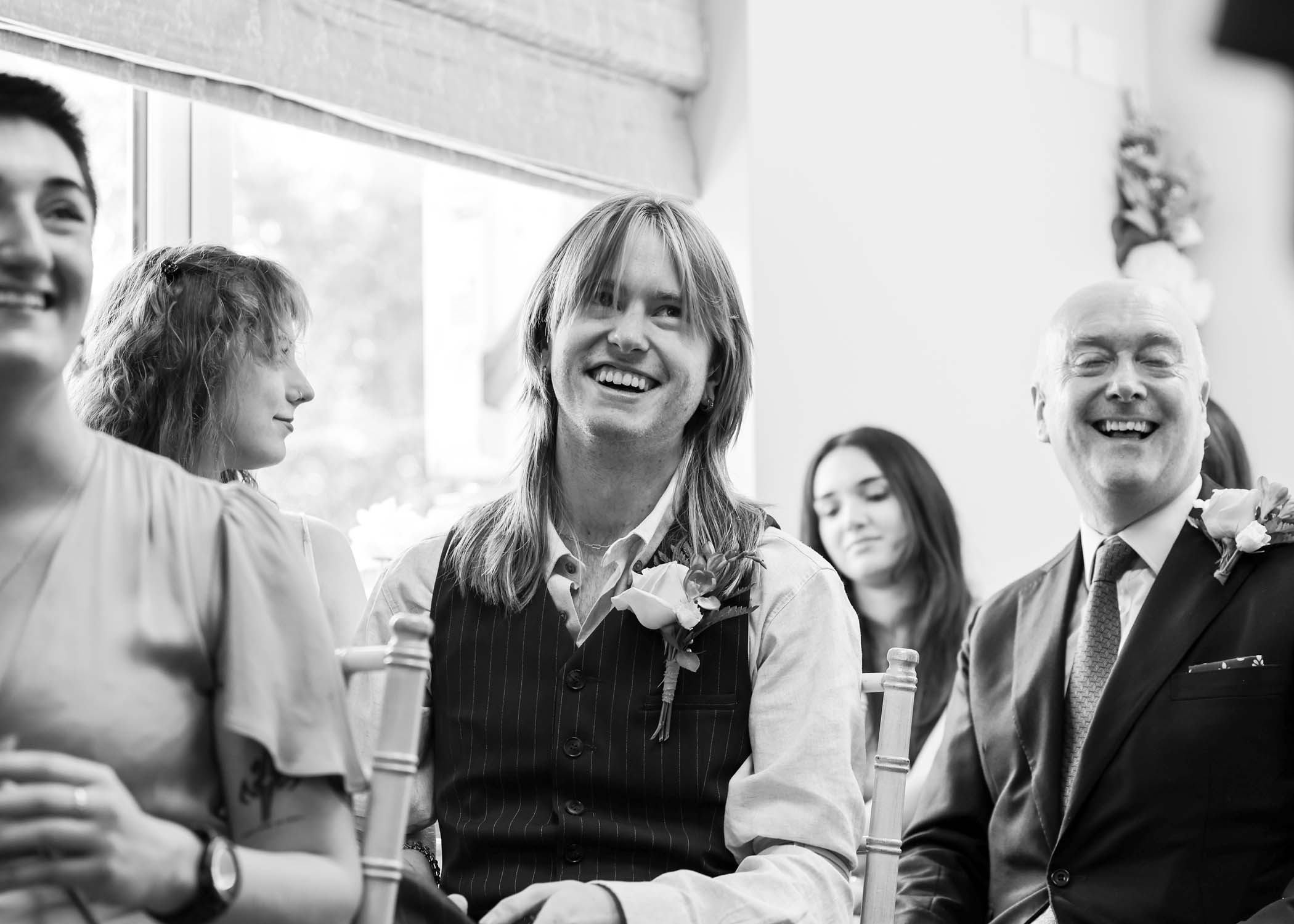 A black and white photo of a wedding guest smiling during the wedding ceremony on a wedding day in Woodbridge captured by Suffolk Wedding Photographer Hayley Denston Photography